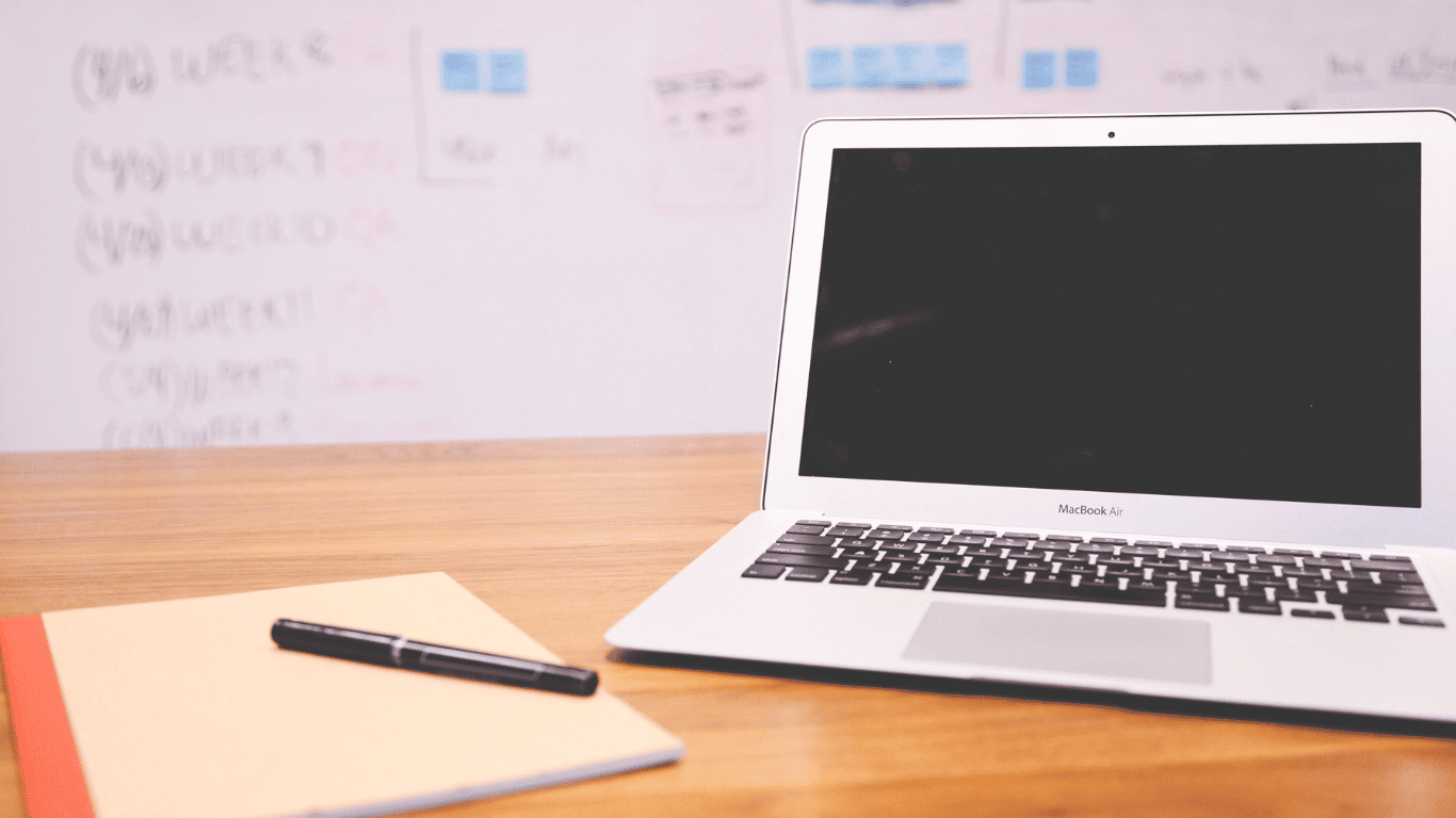 Laptop and notebook with pen on a desk in front of a whiteboard with notes.