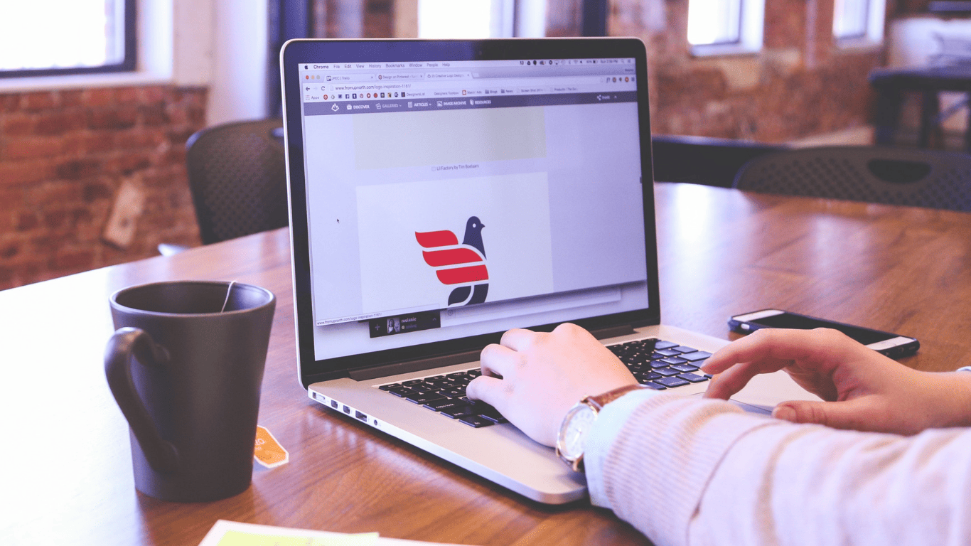 Person working on a laptop designing a logo, with a coffee mug and smartphone on the desk.