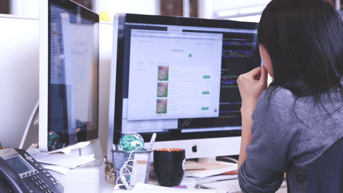 Woman working at a desk with dual computer screens in an office setting.
