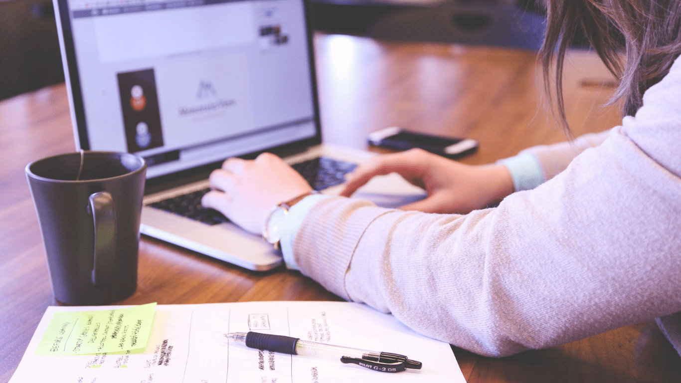 Woman working on a laptop with notes, pen, and coffee mug on the desk.