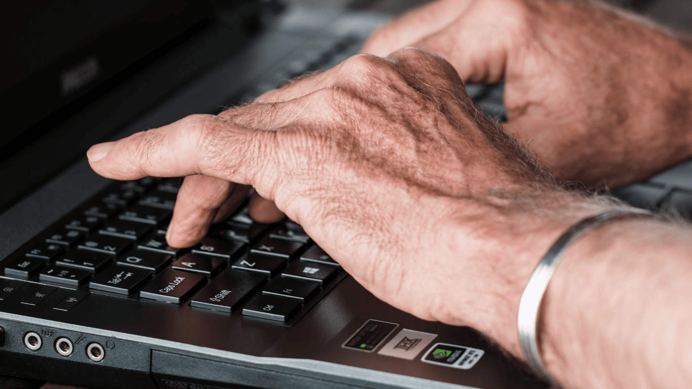 Close-up of elderly hands typing on a laptop keyboard.