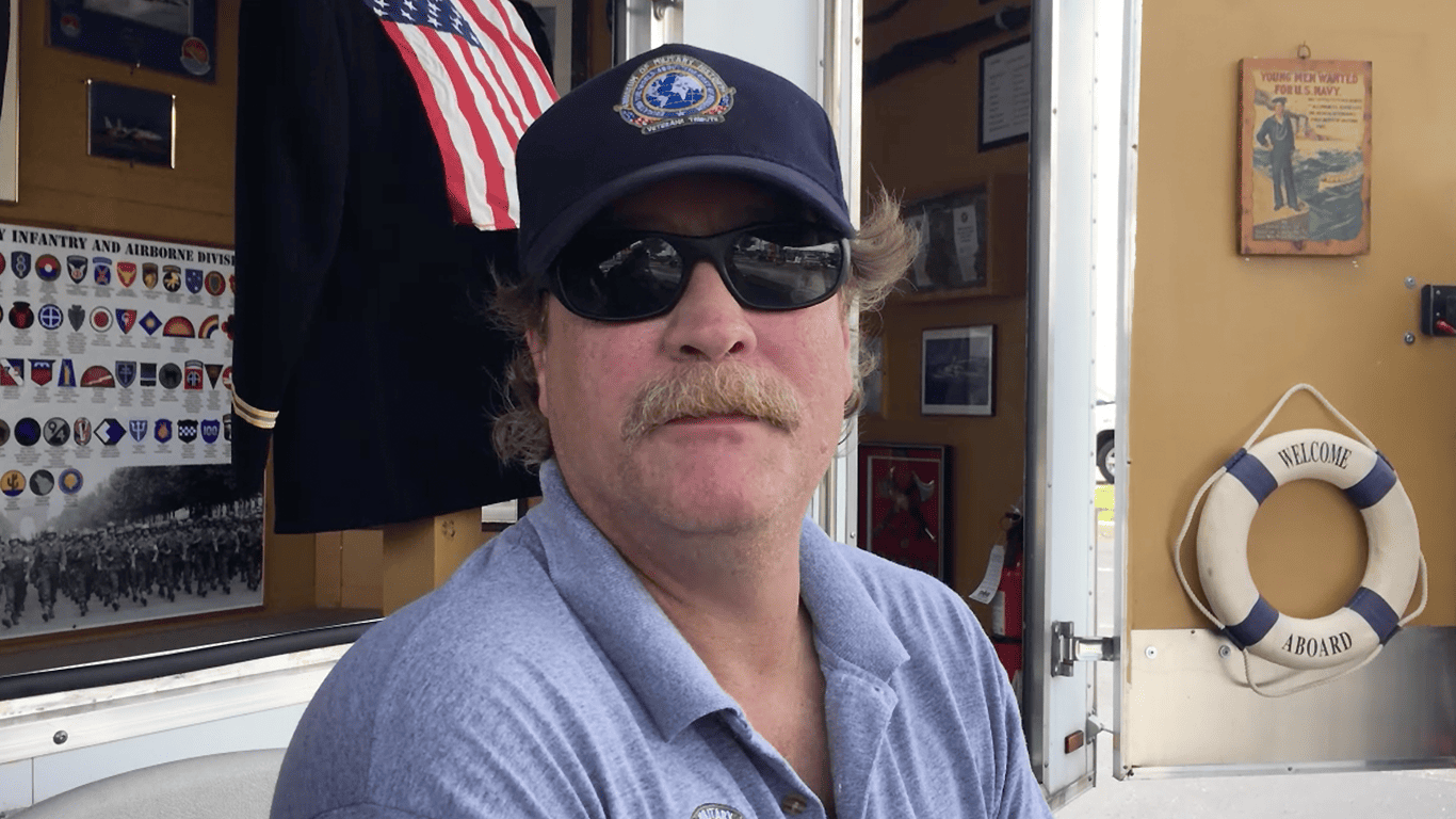 Man wearing sunglasses and a cap sitting in front of a U.S. Navy display with military memorabilia.