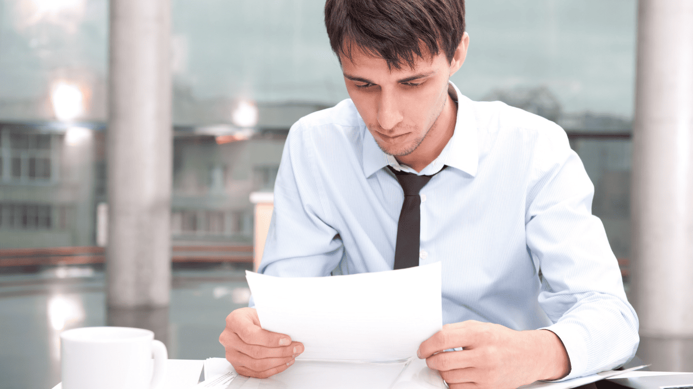 Businessman in formal attire carefully reviewing financial documents at a desk with coffee cup nearby.
