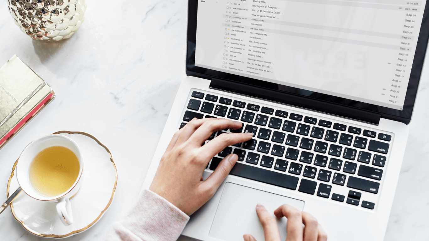 Person typing on a laptop and checking emails with a cup of tea on the desk.