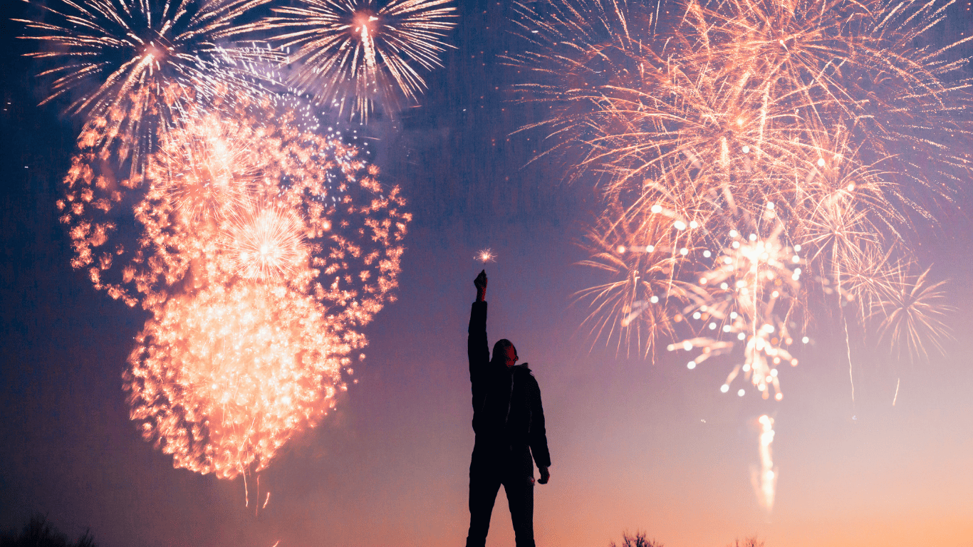 Person holding a sparkler with colorful fireworks lighting up the night sky
