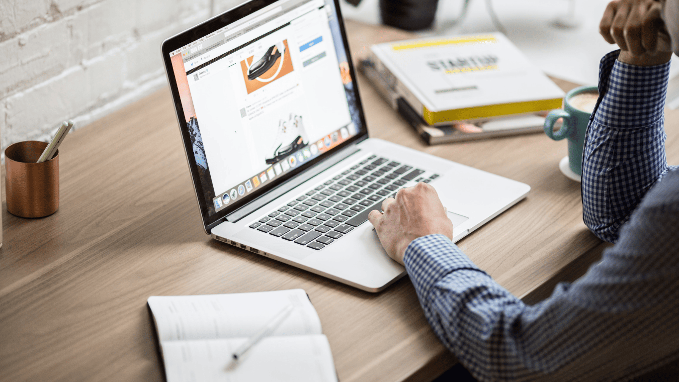 Man browsing online content on a laptop at a desk with books, coffee, and a notebook.