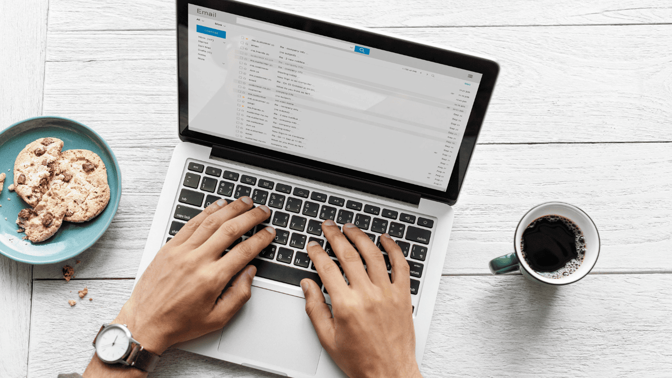 Person typing on a laptop while checking emails with coffee and cookies on the desk.