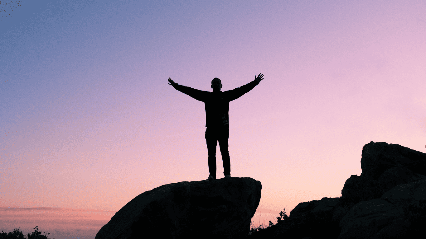 Silhouette of a person standing on a rock with arms raised against a sunset sky, symbolizing success and freedom.