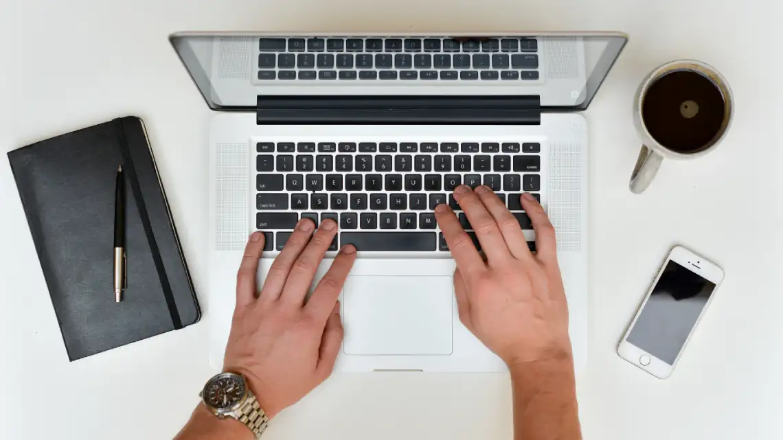 Top view of a person typing on a laptop, with a notebook, pen, smartphone, and a cup of coffee placed nearby on a white desk.