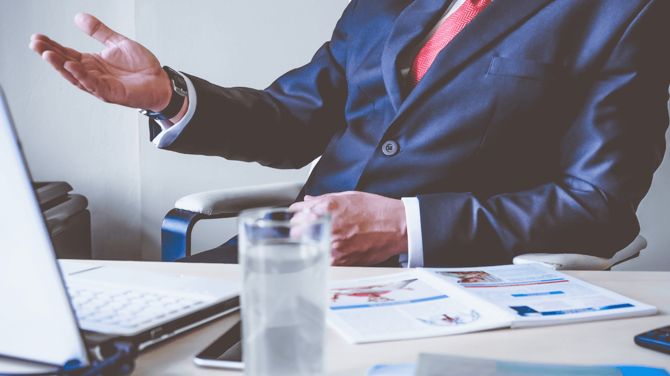 Business professional in a suit gesturing during a meeting or presentation at a desk with documents and a laptop.