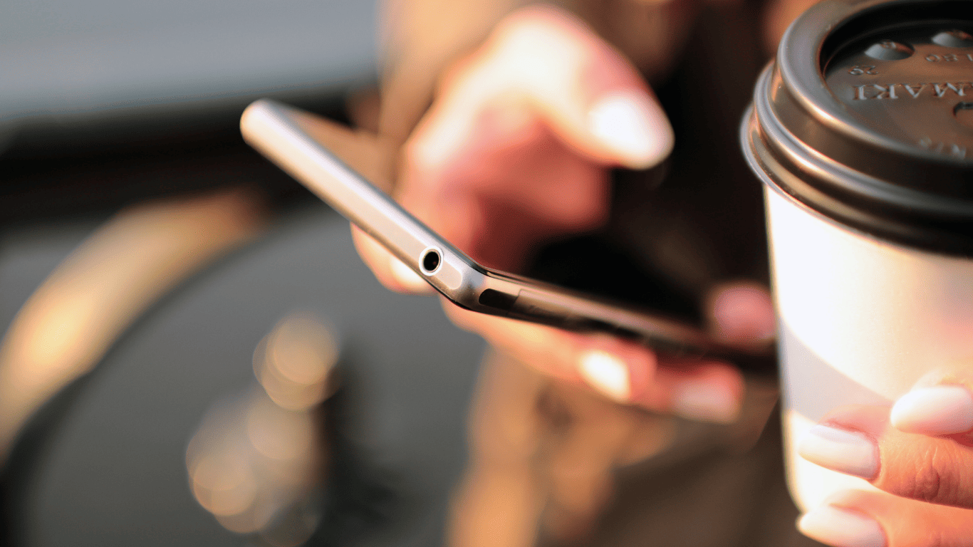 Person holding a smartphone in one hand and a takeaway coffee cup in the other.