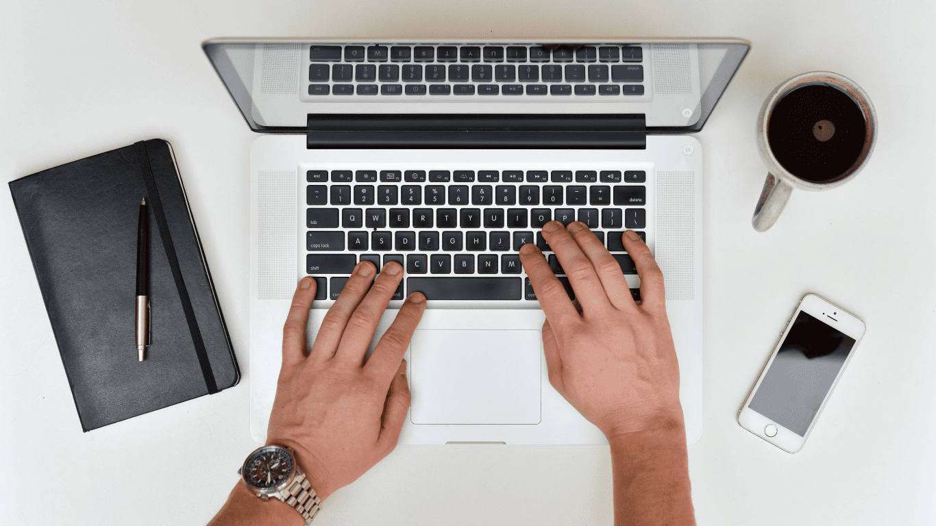 Person typing on a laptop with a notebook, pen, coffee, and smartphone on the desk.