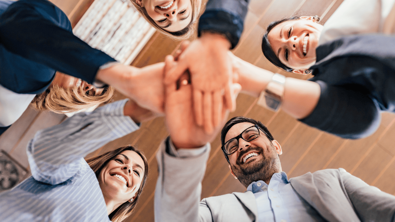 Group of smiling colleagues standing in a circle with hands stacked together in teamwork celebration.