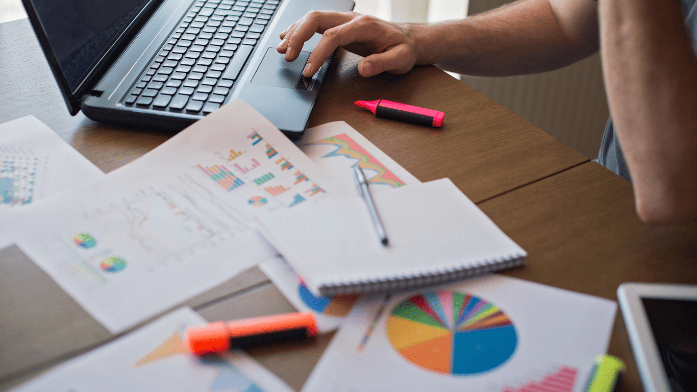 Person working on a laptop with financial charts, reports, and graphs spread across the desk.