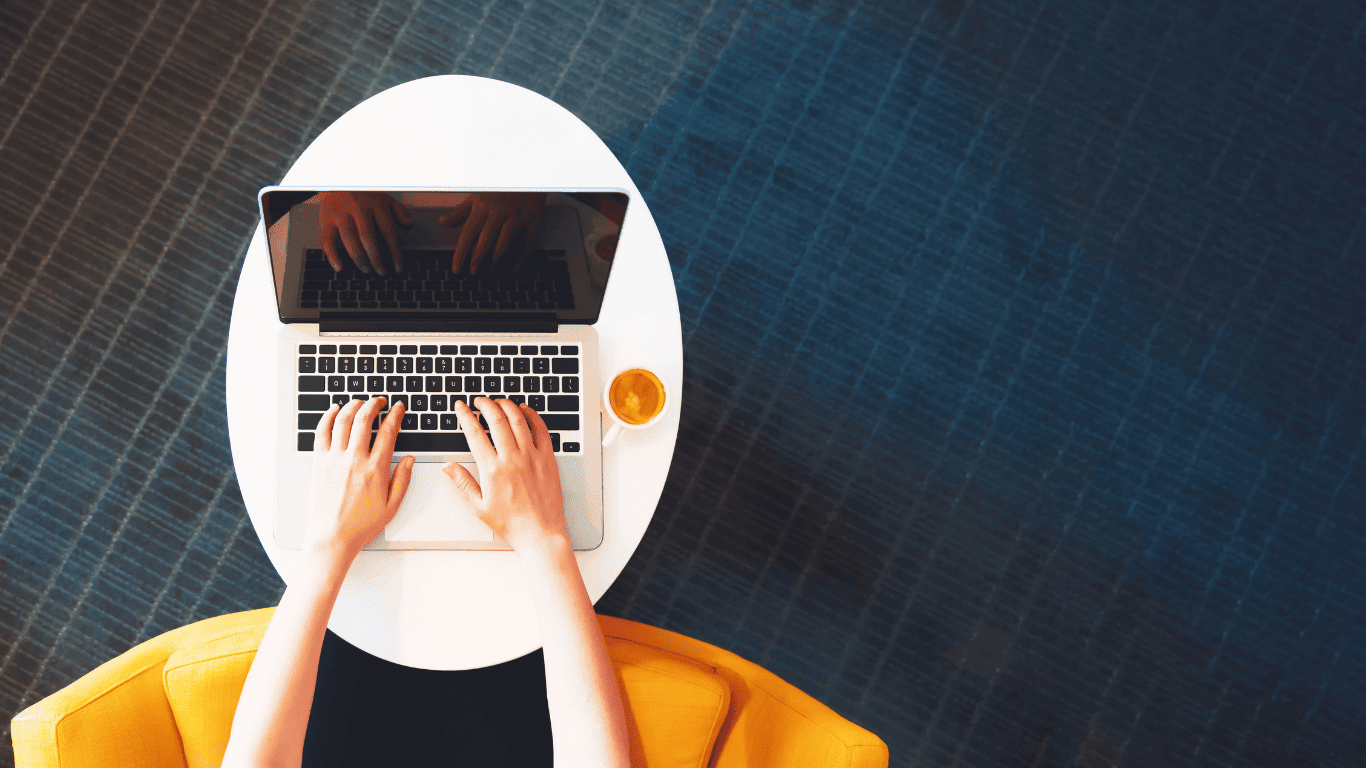 Person typing on a laptop at a round white desk with a cup of coffee.