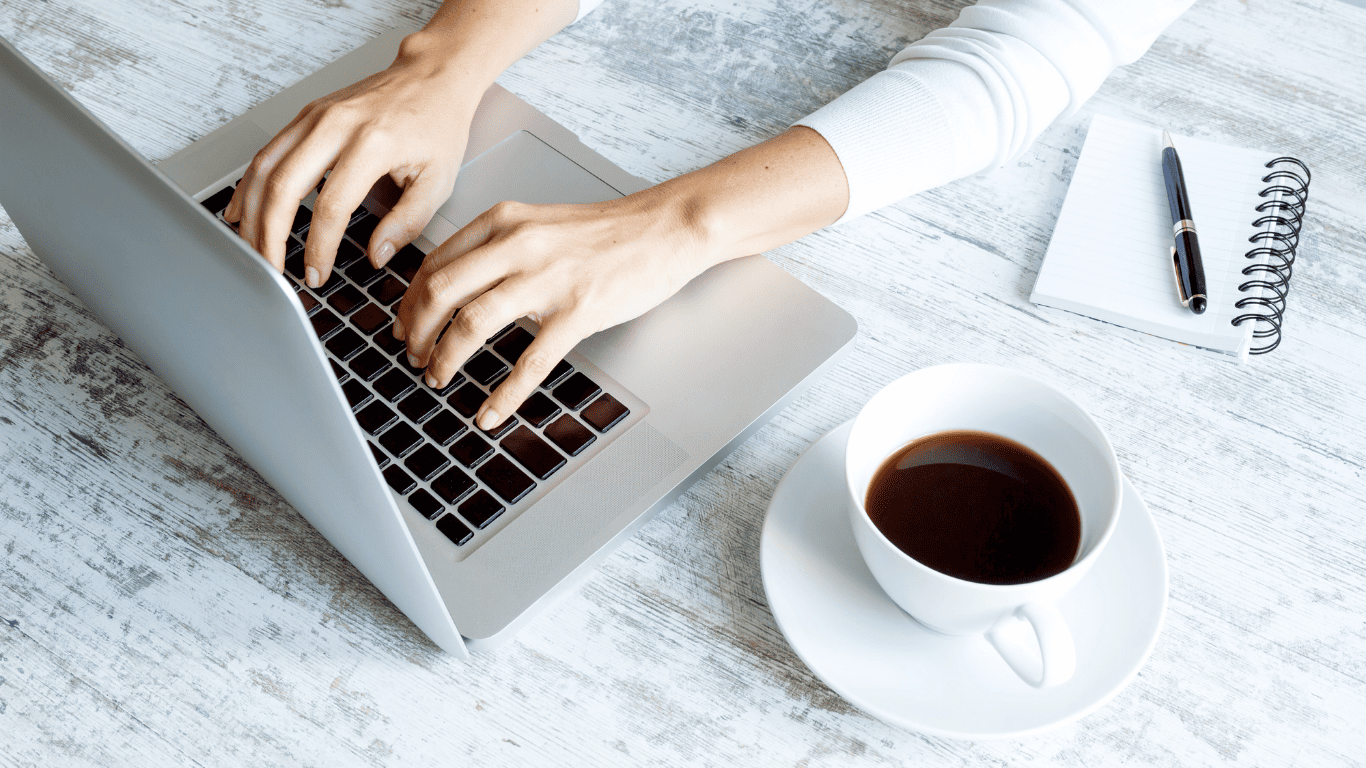 Person typing on a laptop with a cup of coffee and notepad on the desk.